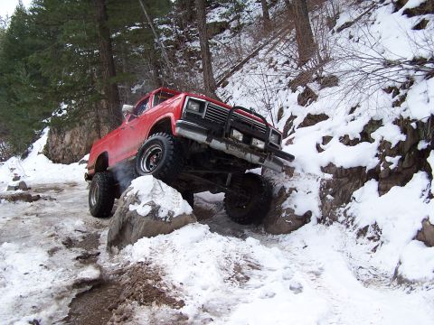 1985 Ford Bronco - This modified truck is shown off-roading on a challenging snowy incline with large rocks.