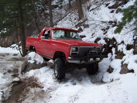 1985 Ford Bronco - This red off-road vehicle is shown navigating a challenging snowy and rocky trail, with a winch visible on its front bumper.