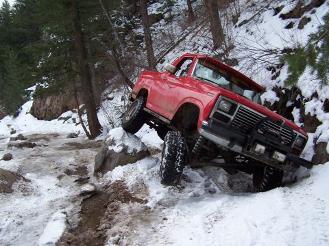 1985 Ford Bronco - Red Ford pickup truck struggling on a snowy, rocky off-road trail.