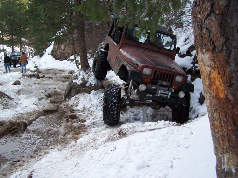 1985 Ford Bronco - A red Jeep Wrangler navigates a challenging, snow-covered trail with large rocks and trees.