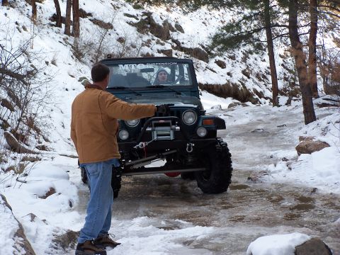 1985 Ford Bronco - A green Jeep Wrangler navigates an icy, snow-covered road in a wooded area, with a person standing outside the vehicle.
