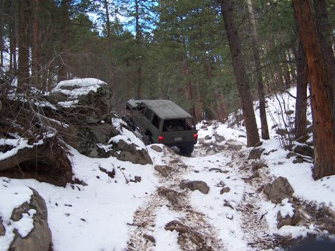 1985 Ford Bronco - An olive green SUV struggles on a snow-covered, rocky path through a dense forest.