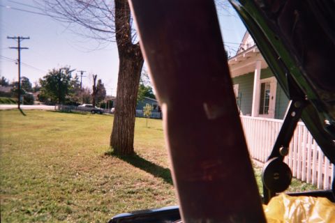 1990 Ford Bronco - grassy front yard with a leafless tree and a green house with white picket fence, framed by a raised vehicle hood.