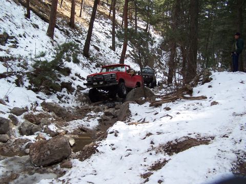 1985 Ford Bronco - Two off-road vehicles tackle a challenging snowy and rocky path through a wooded area.