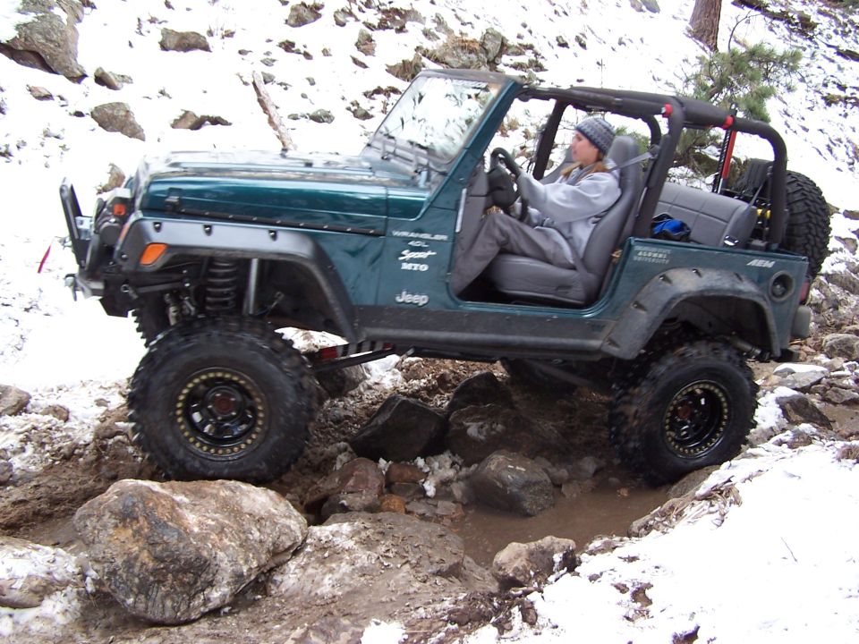 Green Jeep Wrangler driving over rocks and through mud with snow on the ground. Driver is wearing a beanie and jacket