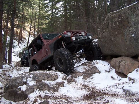 1985 Ford Bronco - A maroon Jeep Wrangler navigates a challenging, snow-dusted rocky terrain with another vehicle visible behind it.