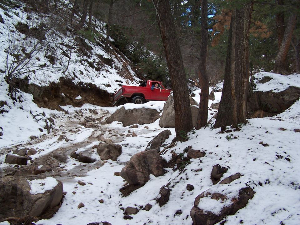 Red pickup truck navigating a snowy, rocky off-road trail with trees on either side