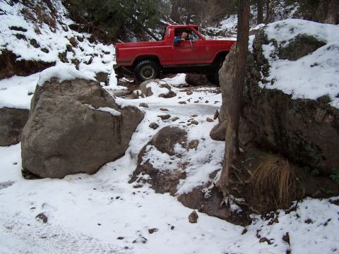 1985 Ford Bronco - A red pickup truck drives through a snowy, rocky terrain, with ice visible on the ground.