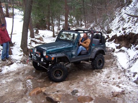 1985 Ford Bronco - A green Jeep Wrangler navigates a challenging off-road path covered in snow and mud.