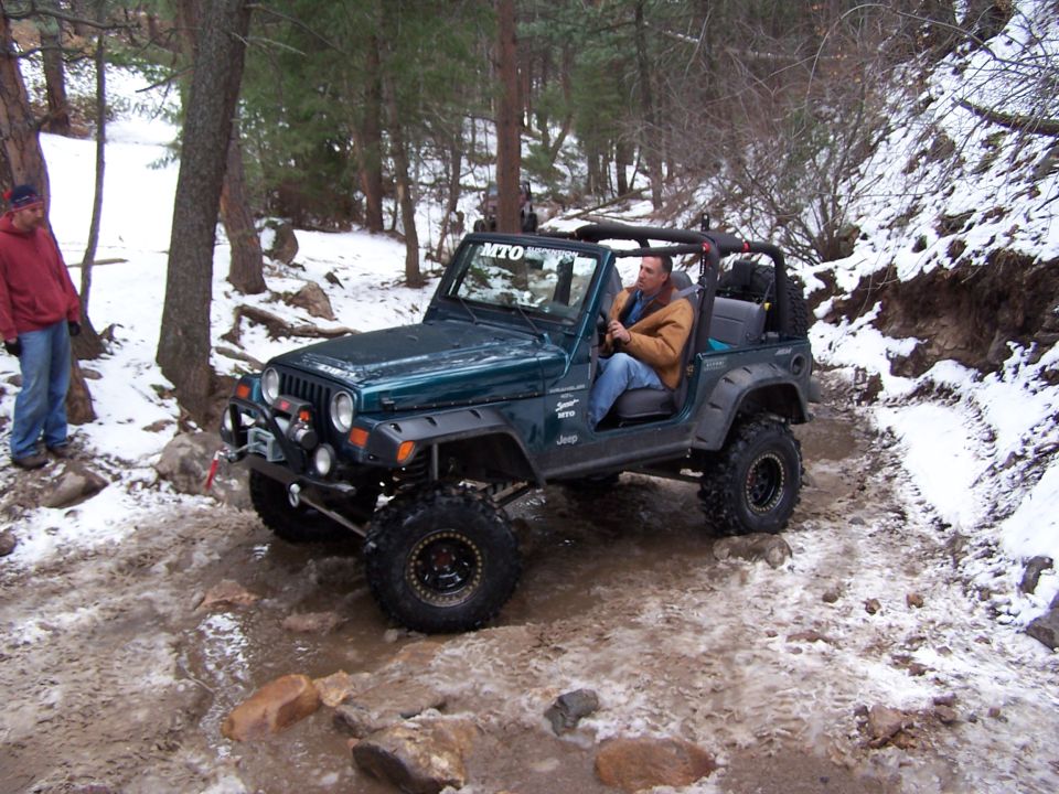Green Jeep Wrangler driving on a snowy, rocky trail with a man at the wheel