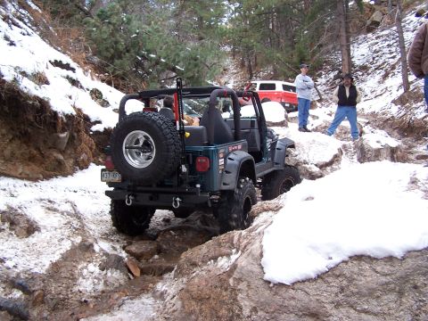 1985 Ford Bronco - A dark green Jeep Wrangler tackles a challenging snowy and rocky path, with a red SUV visible further up the trail.
