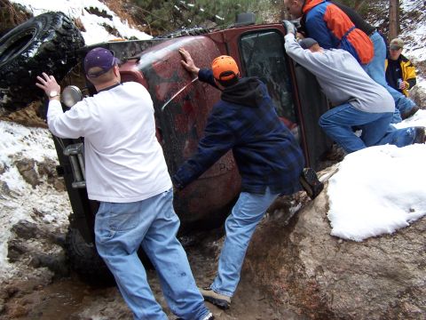 1985 Ford Bronco - Five people push against the side of a maroon 4x4 rolled onto its side in a snowy, rocky trail gully, working to right it back onto its whe…