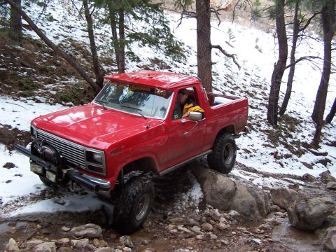 1985 Ford Bronco - This lifted pickup truck navigates a challenging rocky terrain with snow patches on the ground.