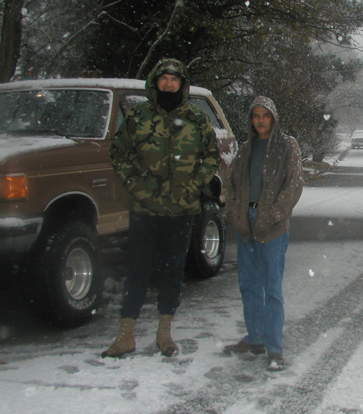 Two men in winter coats stand by a brown 1987 Ford Bronco in the snow. The Bronco has aftermarket wheels