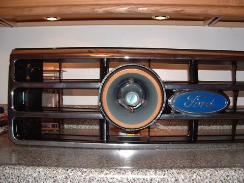 1990 Ford Bronco - A chrome-and-black horizontal-bar Ford truck grille standing upright on a granite countertop with a car audio speaker mounted through the r…