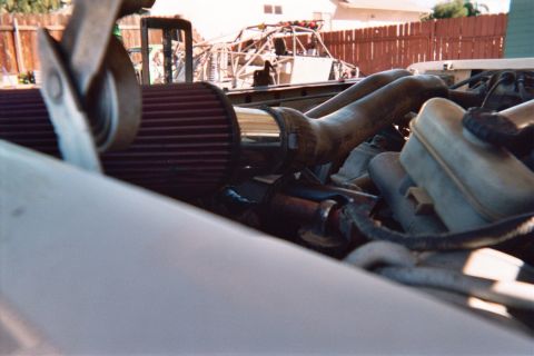 1990 Ford Bronco - Close engine-bay view of a red cone air filter and intake tubing routed past the brake reservoir, fence behind.