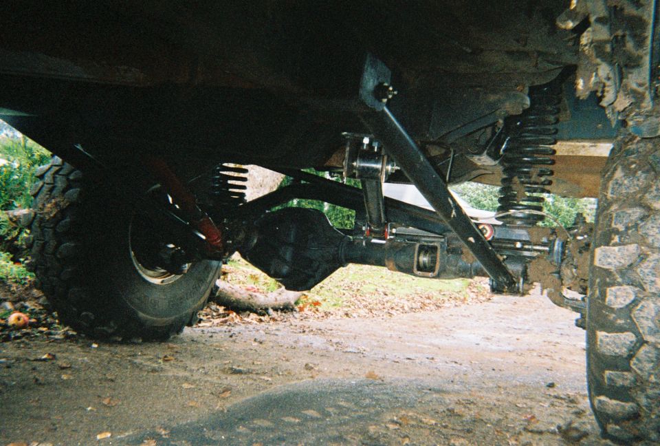 Underside of lifted 4x4 showing front axle, coil spring, control links and off-road tires.