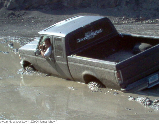 Mud-covered extended-cab Ford pickup stuck door-deep in a muddy water hole