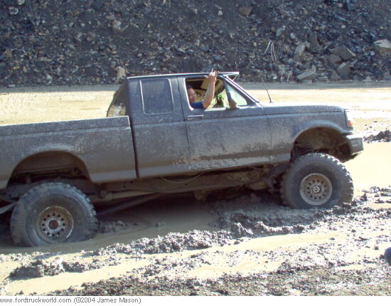 Lifted extended-cab pickup covered in mud driving through a mud pit, driver leaning out window