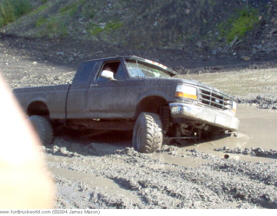 Mud-covered 90s Ford F-150 extended cab stuck axle-deep in a mud pit on mud tires