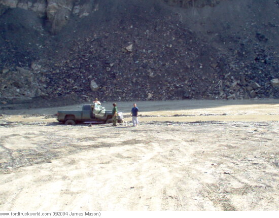 Tan single-cab pickup parked at the base of a rock quarry with two people beside it