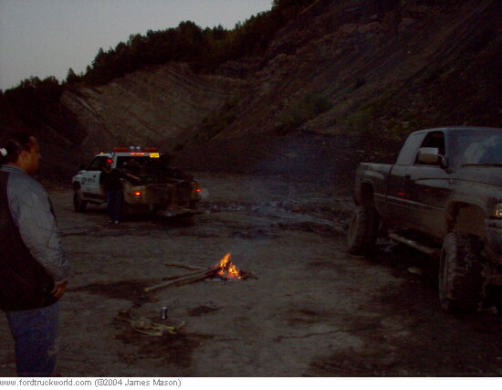 Lifted pickup on mud tires beside a campfire in a gravel pit at dusk
