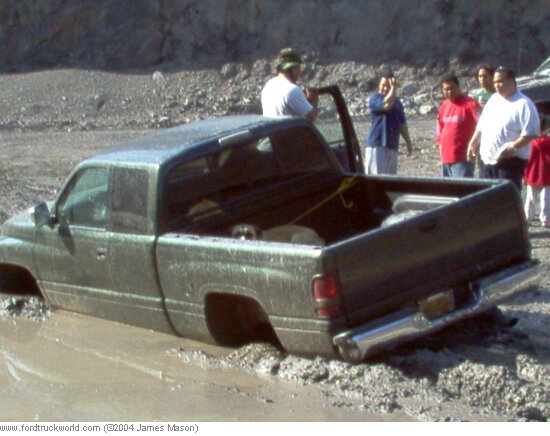 Green Dodge Ram pickup stuck to its rockers in a muddy pit with onlookers