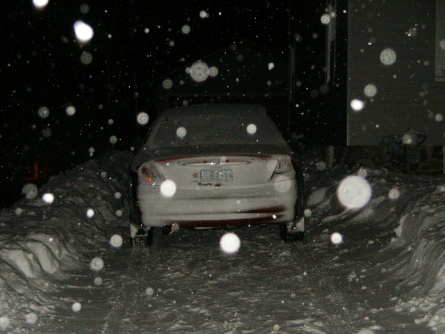 Rear view of a white 2000 Ford Taurus covered in snow, parked in a snowy driveway at night