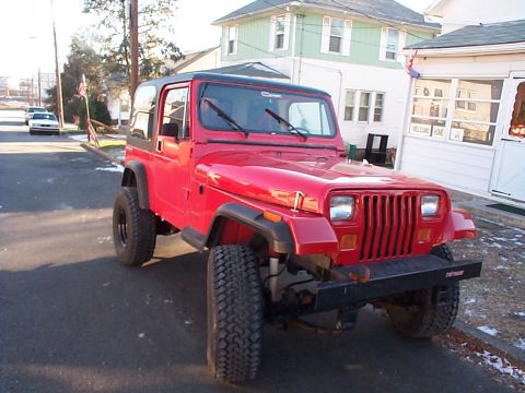 1989 Jeep YJ - A red Jeep Wrangler is parked on a street in front of houses on a sunny day.