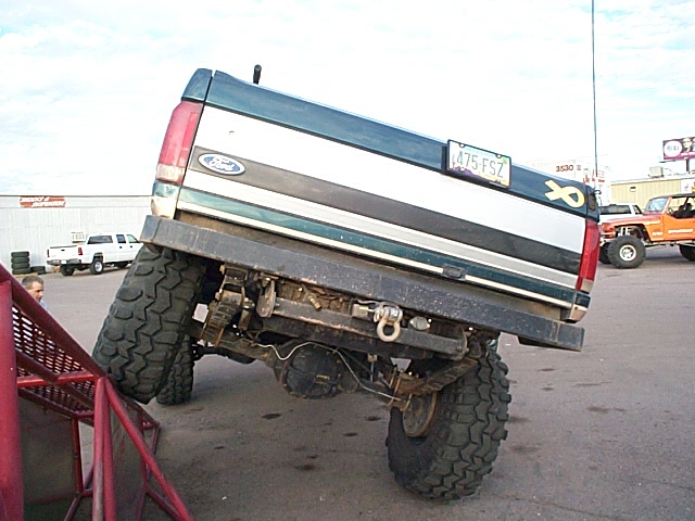 Rear of green/silver lifted Ford truck with front end up, showing axle and big mud tires