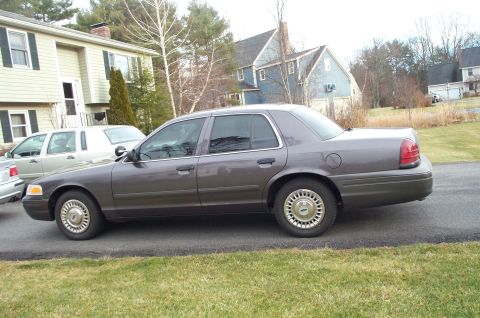 2001 Ford Crown Victoria - A gray 2001 Ford Crown Victoria is parked on an asphalt driveway next to a house, with another vehicle to its left.
