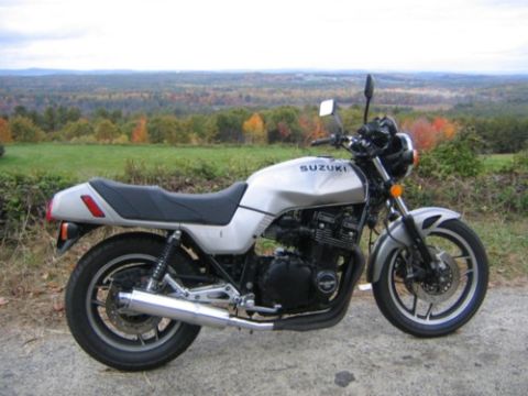 1985 Ford LTD Crown Victoria - A silver Suzuki motorcycle is parked on a dirt road, with a scenic view of rolling hills and autumn trees in the background.