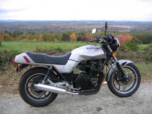 Silver Suzuki motorcycle parked on a dirt road with autumn foliage in the background