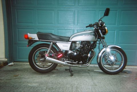 1985 Ford LTD Crown Victoria - A Suzuki GS550 motorcycle is parked on a concrete surface in front of a teal garage door.