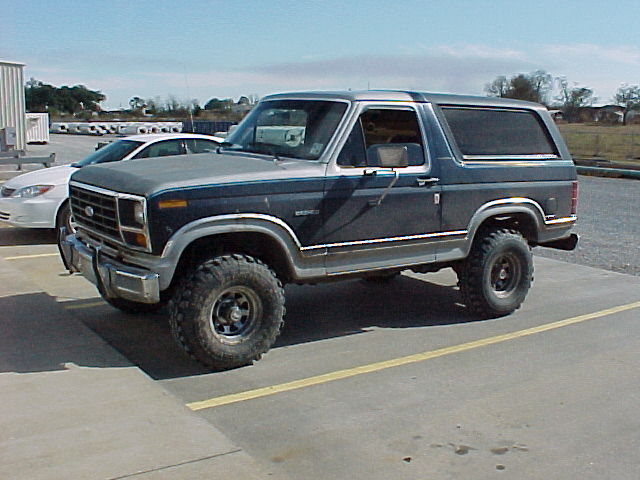 Lifted dark blue Ford Bronco SUV with large off-road tires, driver-side front 3/4 view in a parking lot