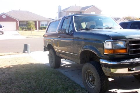 1992 Ford Bronco - Front three-quarter view of a dark green Ford Bronco with a white hardtop, splattered with dried mud down its flanks and wheel arches after…