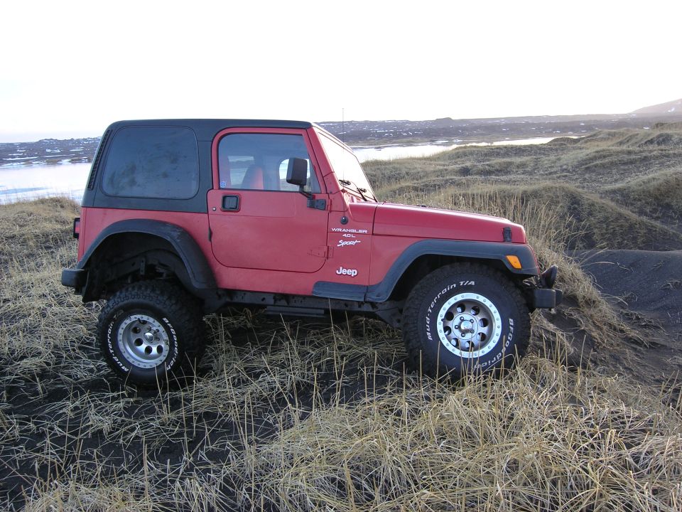 Red Jeep Wrangler with black hardtop and BFGoodrich Mud-Terrain tires parked on grassy black-sand dunes