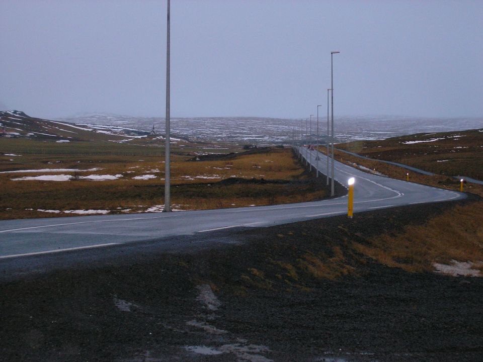 Curving paved road with light poles cutting through a snow-patched plateau at dusk