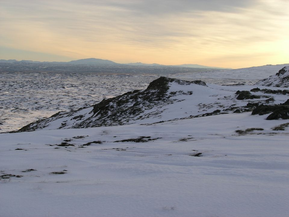 Snow-covered ridgeline with rocky outcrop overlooking distant hills at sunset