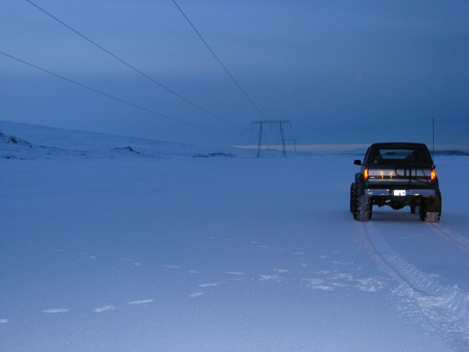 Lifted pickup with brake lights on, driving away across a snowy plain at dusk under power lines
