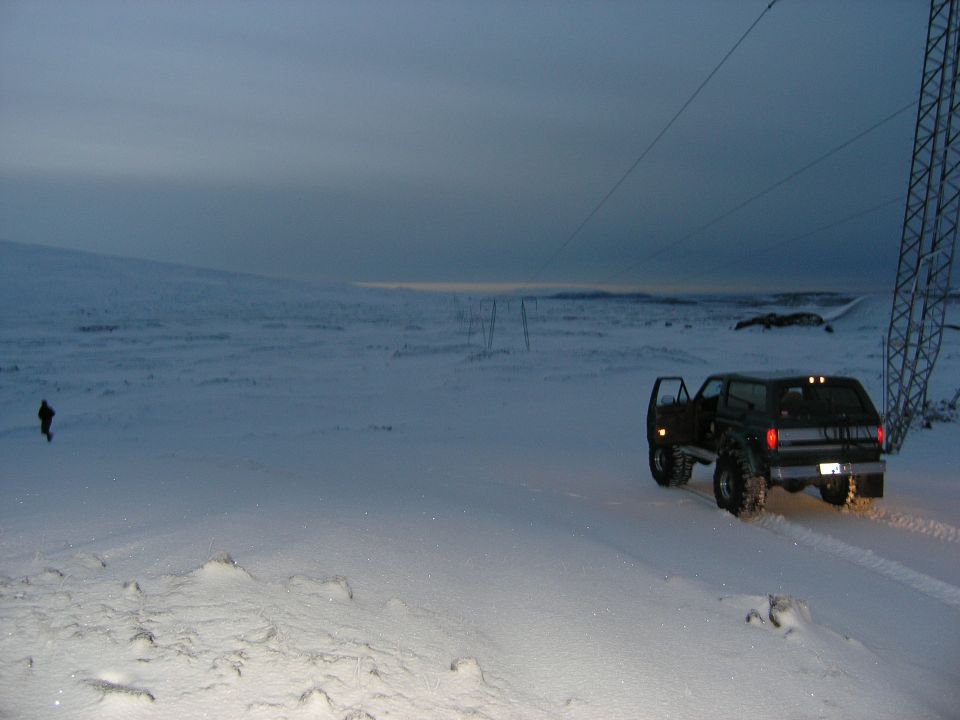 Lifted dark SUV with driver door open on a snowy slope under power lines at dusk, person walking nearby