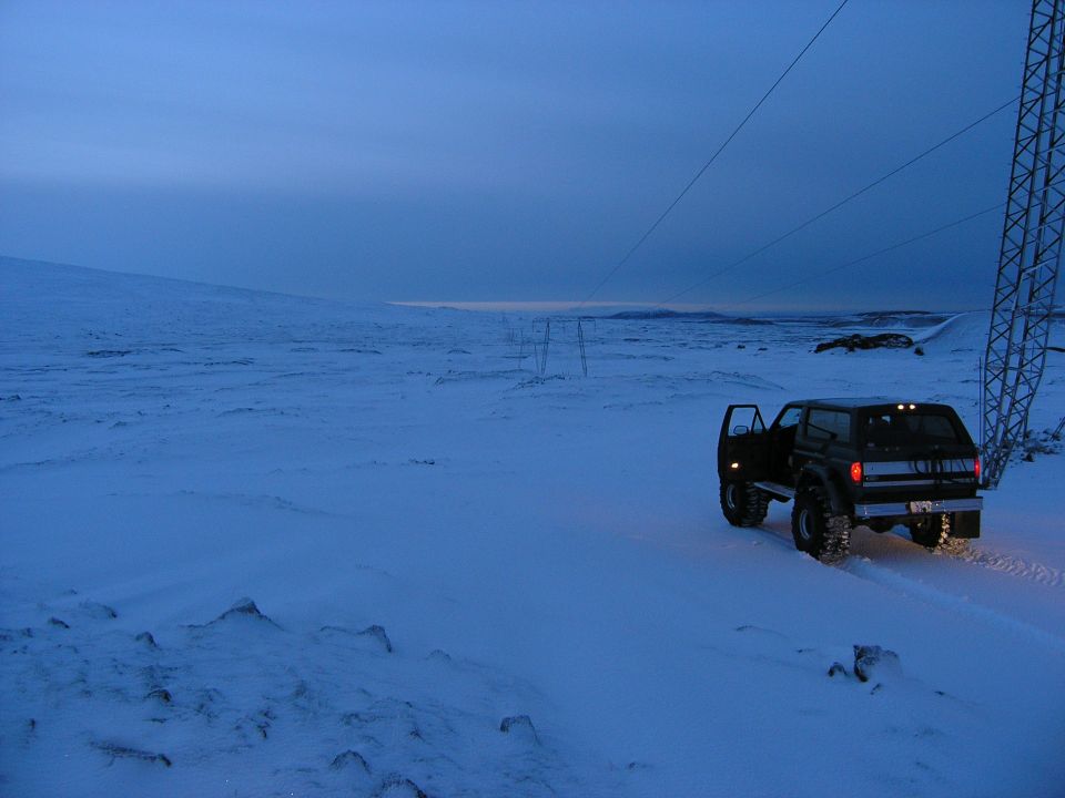 Lifted dark SUV with open door parked on a vast snowfield at blue-hour under a power-line tower