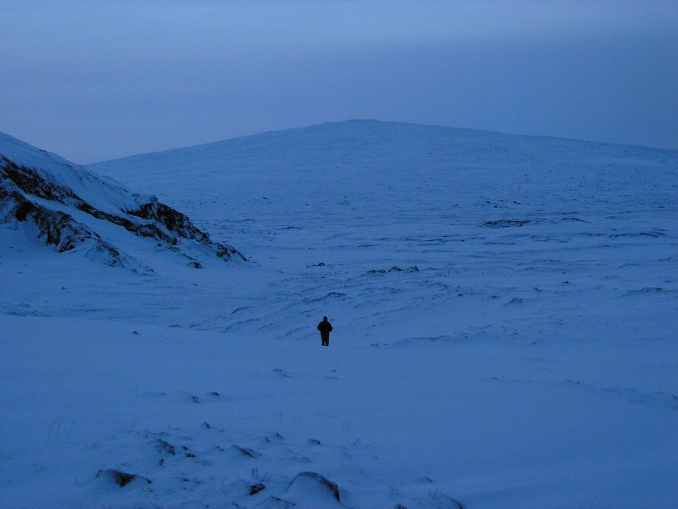 Lone figure standing in a vast snowy basin below a rounded hill at twilight