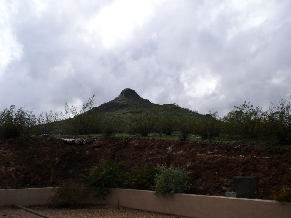 Mountain peak under cloudy sky, with scrub brush and dirt in foreground