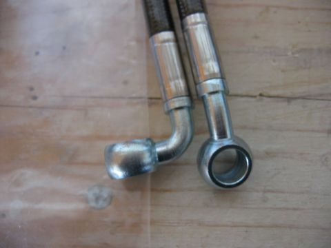 1985 Ford LTD Crown Victoria - Two braided brake lines with metal fittings are shown against a wooden background.