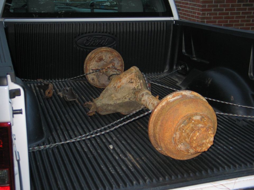 Rusty rear axle with brake drums roped down in the black bed liner of a white Ford pickup