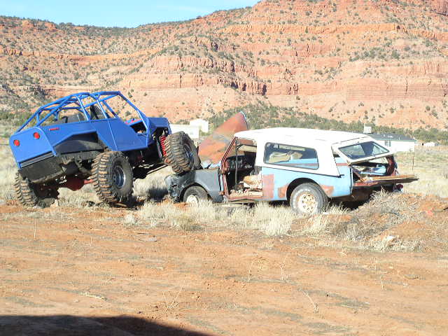 Blue off-road buggy on top of a wrecked white and blue SUV in a desert landscape
