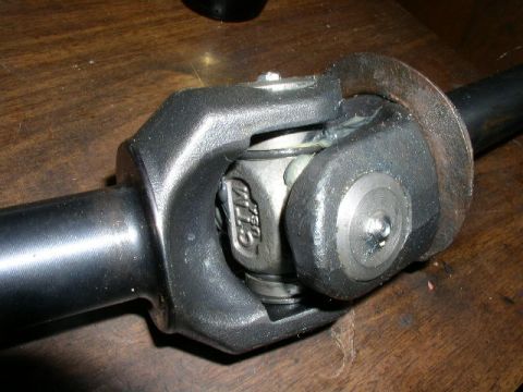 1978 Ford Bronco - Close-up of a driveshaft universal joint laid on a wooden bench, showing the two black yokes and the bare-metal U-joint cross with cast let…