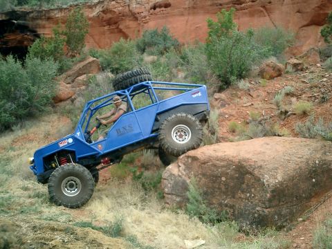 1979 Ford Bronco - Custom buggy tackles a large rock obstacle on a trail. The vehicle has a full roll cage and spare tire mounted on top.