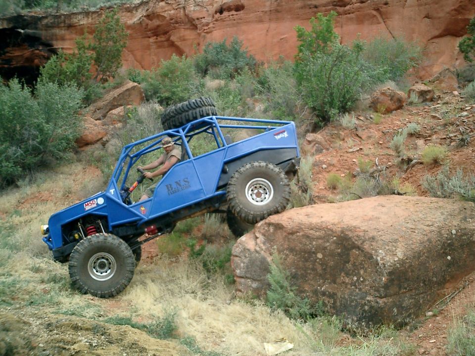 Blue tube-frame rock buggy with roof spare tire crawling over red sandstone boulders